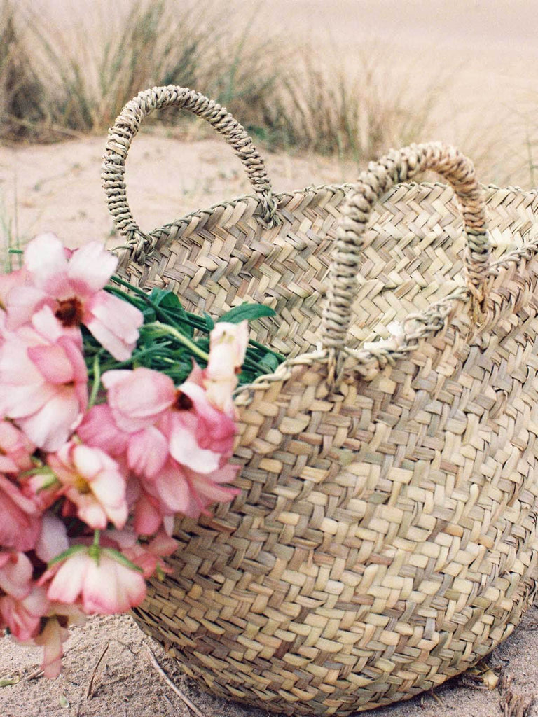 Close up of the natural, rustic Bohemia Beldi Basket filled with pink flowers on a beach