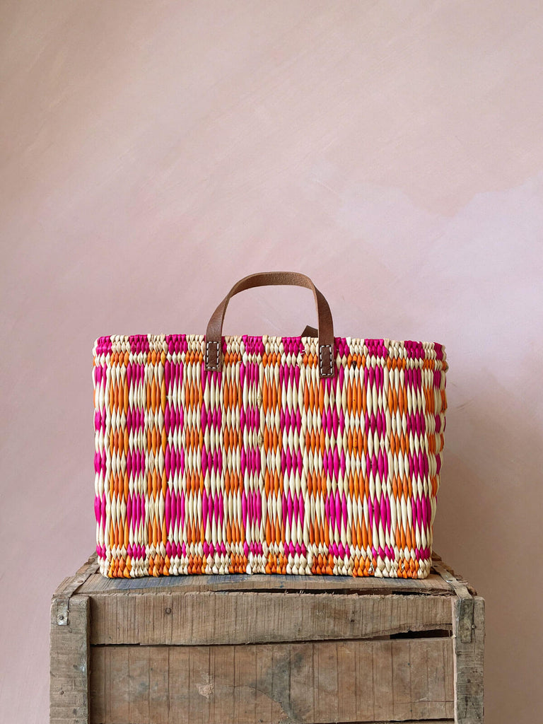 A medium, rectangular, pink and orange chequered woven basket bag with leather handles sitting on a wooden crate