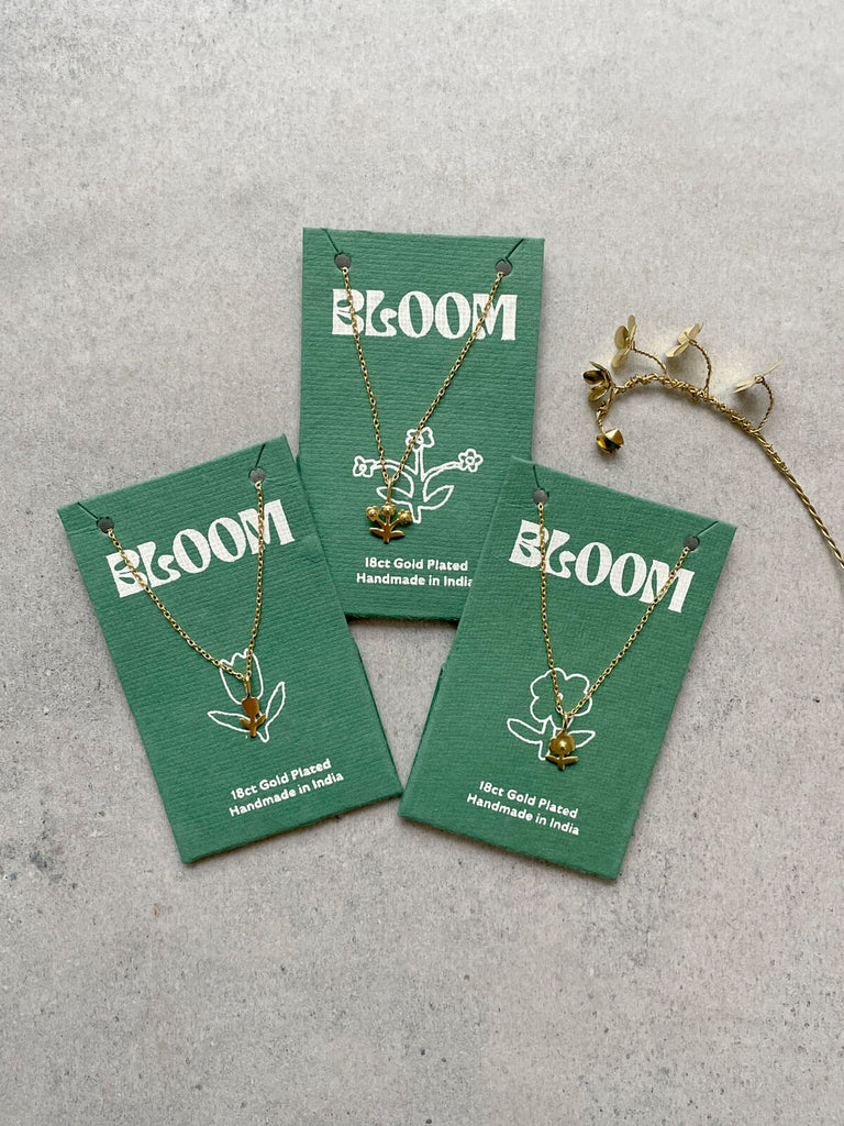 Three Bloom collection gold necklaces on green cotton paper packaging with delicate flower illustrations