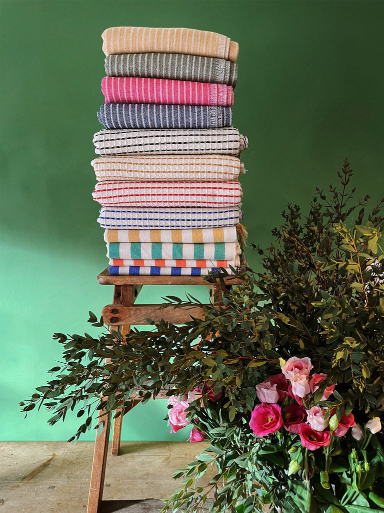 A display of Turkish hammam towels featuring a range of colours, patterns, and textures, neatly folded on a rustic wooden stool against a deep green backdrop | Bohemia Design