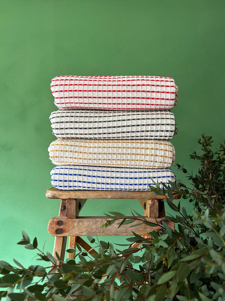 A stack of Milos Hammam Towels in four distinct colours, highlighting the beautiful soft waffle texture of the cotton fabric against a backdrop of lush green wall