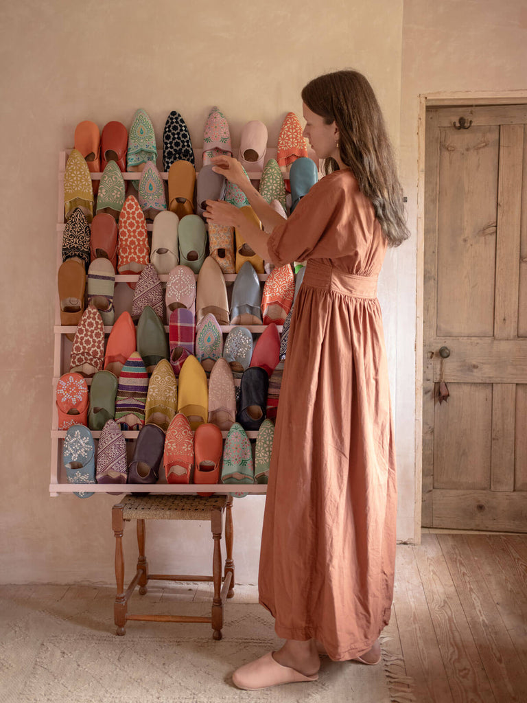 A women in a peach dress wearing a pair of ballet pink Moroccan babouche slippers next to a display of many different styles of Bohemia slippers
