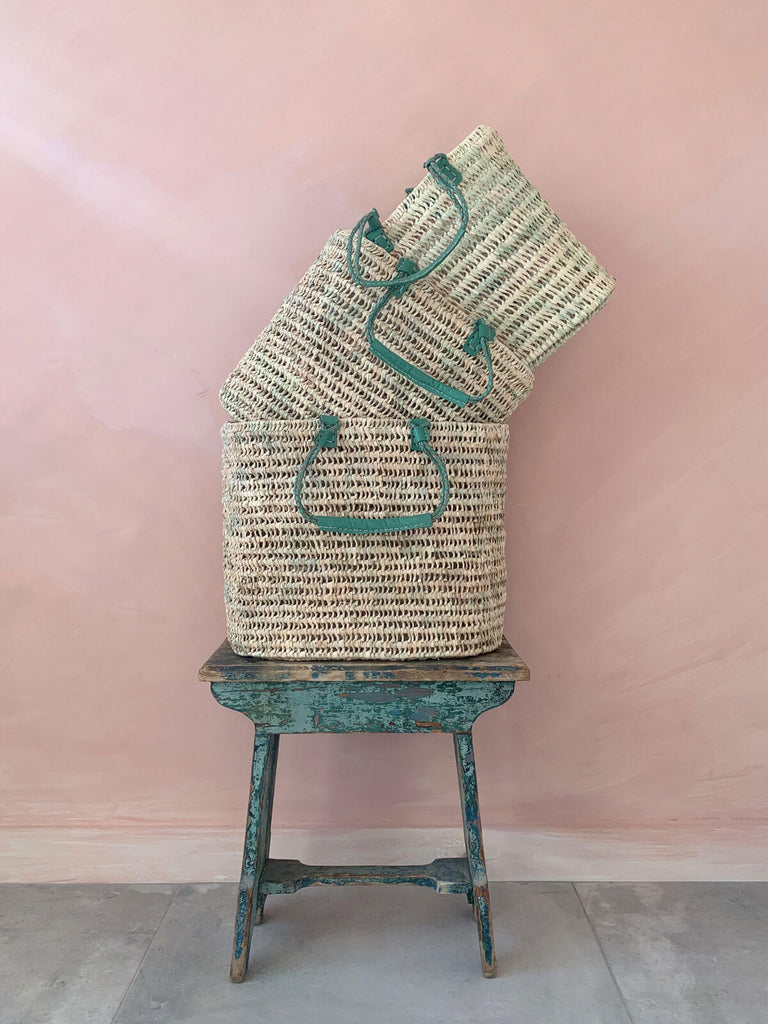 Three sage pleated leather handle baskets on a blue vintage side table in front of a pink wall
