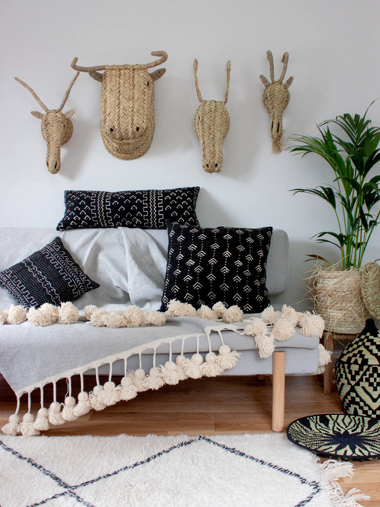 A living room with black mudcloth cushions, a grey pom-pom blanket and plants styled with woven animal heads by Bohemia Design