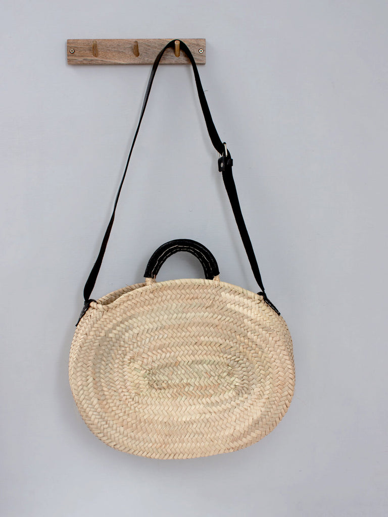 A oval handwoven natural basket bag with black leather straps and two short leather handles by Bohemia Design