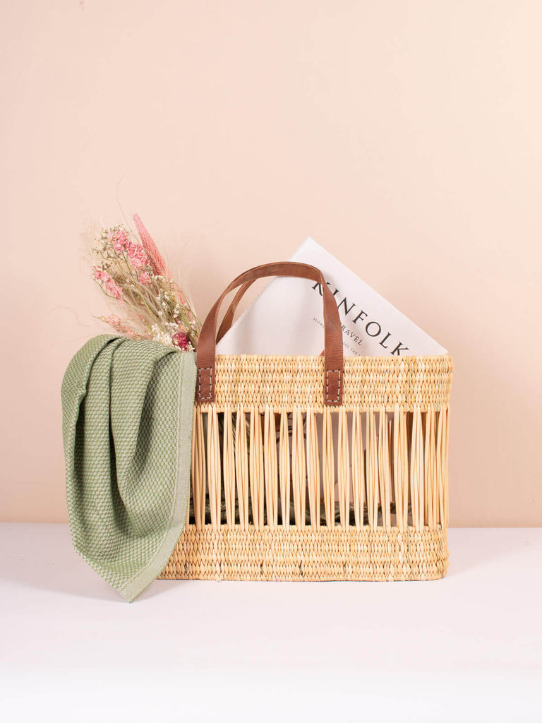 Decorative reed basket with leather handles holding a bunch of dried flowers, scarf and magazine