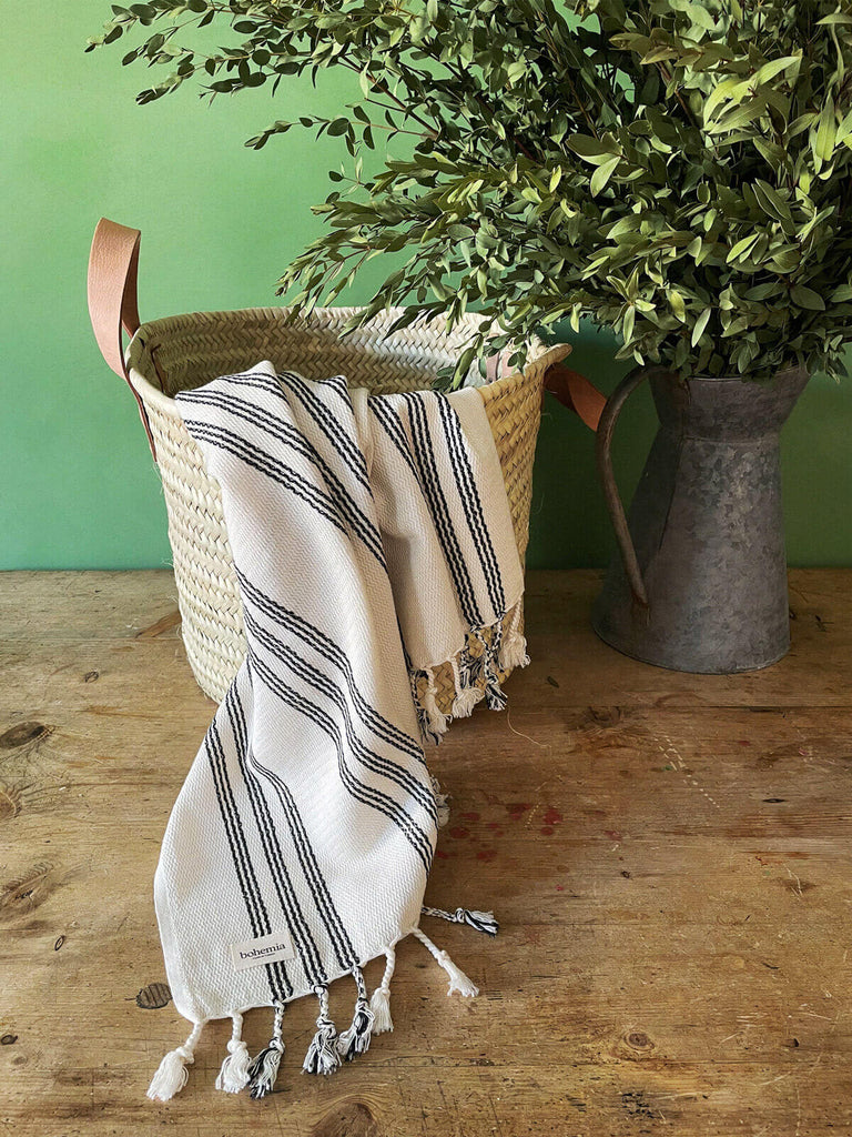 Beautiful Ankara cotton hammam towel with a charcoal ticking stripe, loosely arranged in a natural basket on a wooden floor against a vibrant green wall