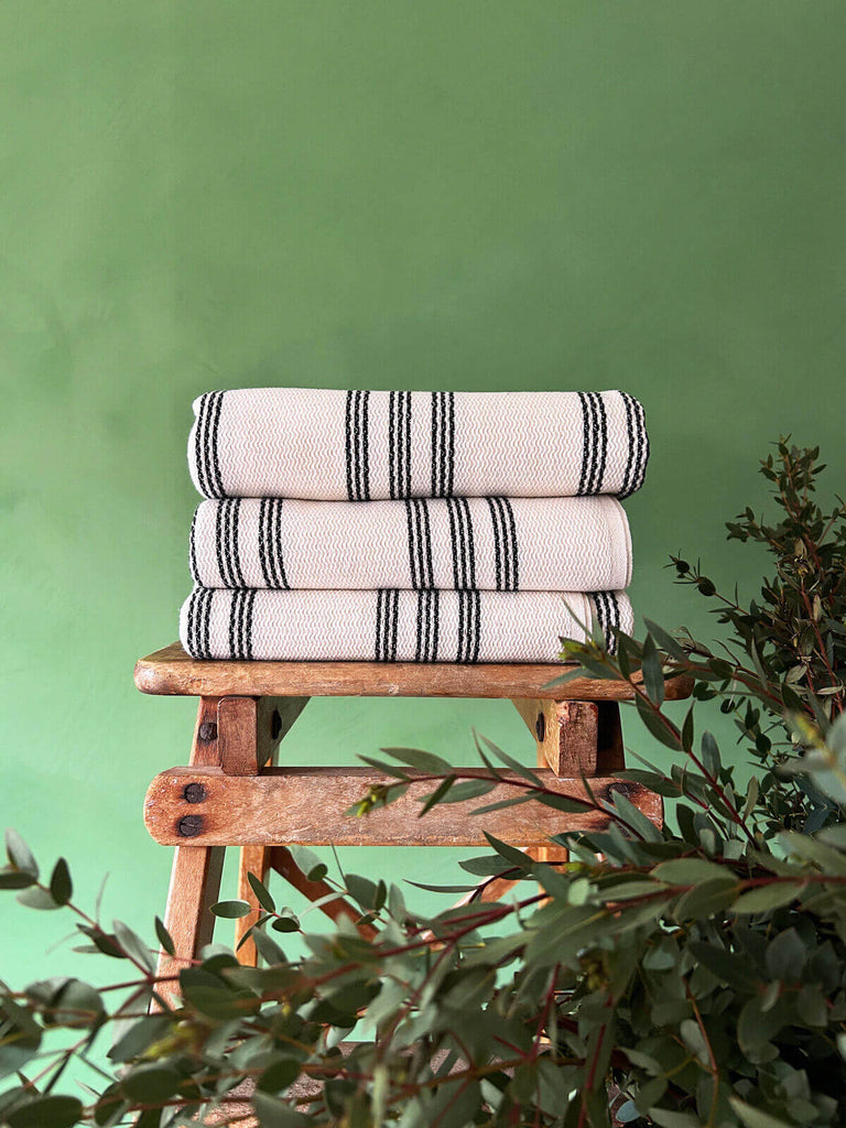 Stack of Turkish hammam towels, showcasing the soft cotton texture and a contrasting charcoal stripe, neatly folded on a wooden stool