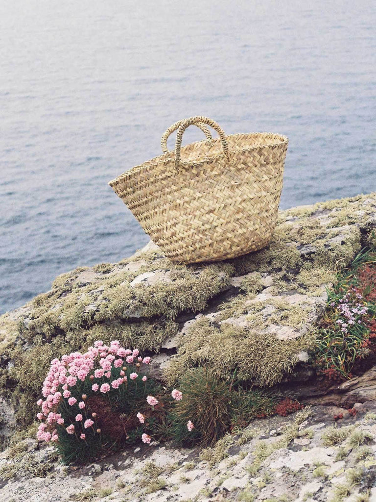 Small natural woven Beldi Basket bag on flower covered rocks by the sea