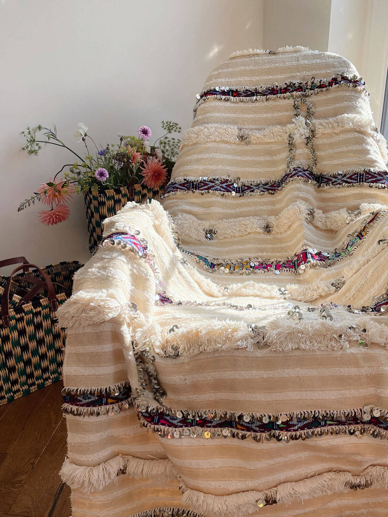 A traditional Moroccan Wedding Blanket draped on a chair features cream and white stripes, beautiful brightly coloured rows of kilim textile, soft fringing and silver sequins