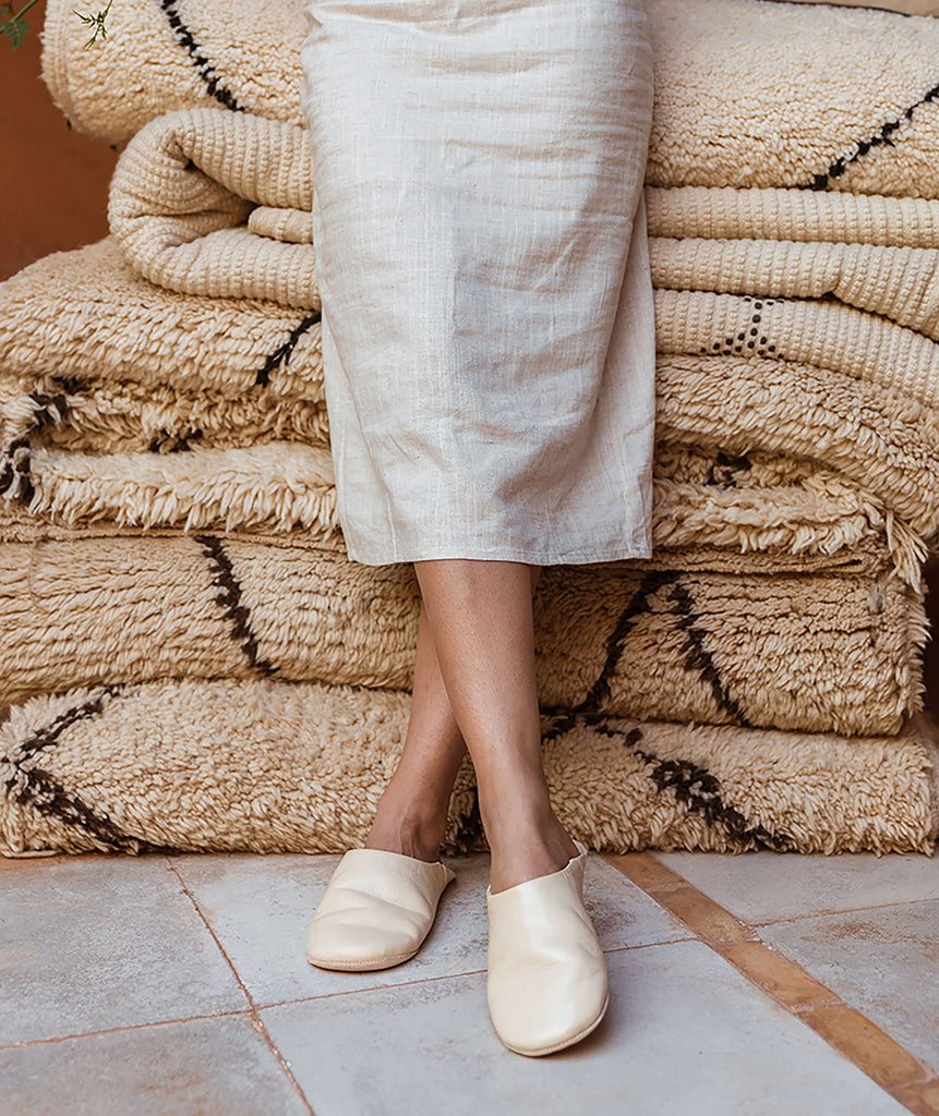 Woman standing against a stack of berber rugs wearing chalk coloured Moroccan babouche slippers by Bohemia design
