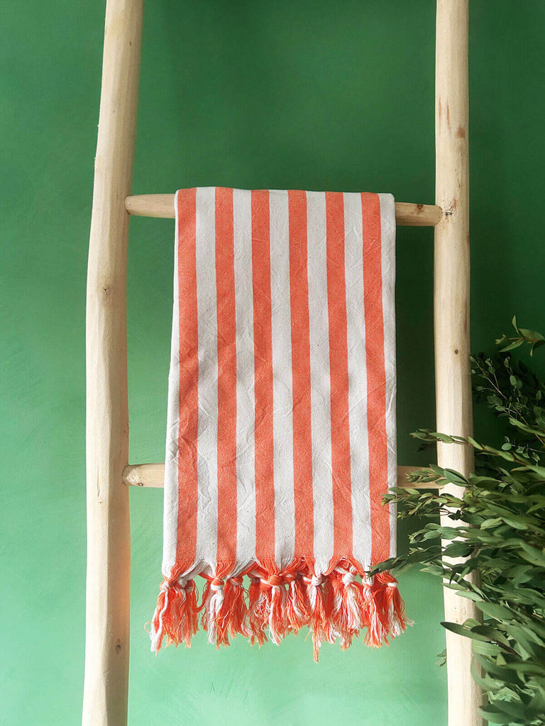 Brighton Stripe cotton hammam towel with a hot orange stripe and fringe, casually displayed on a rustic wooden ladder | Bohemia design