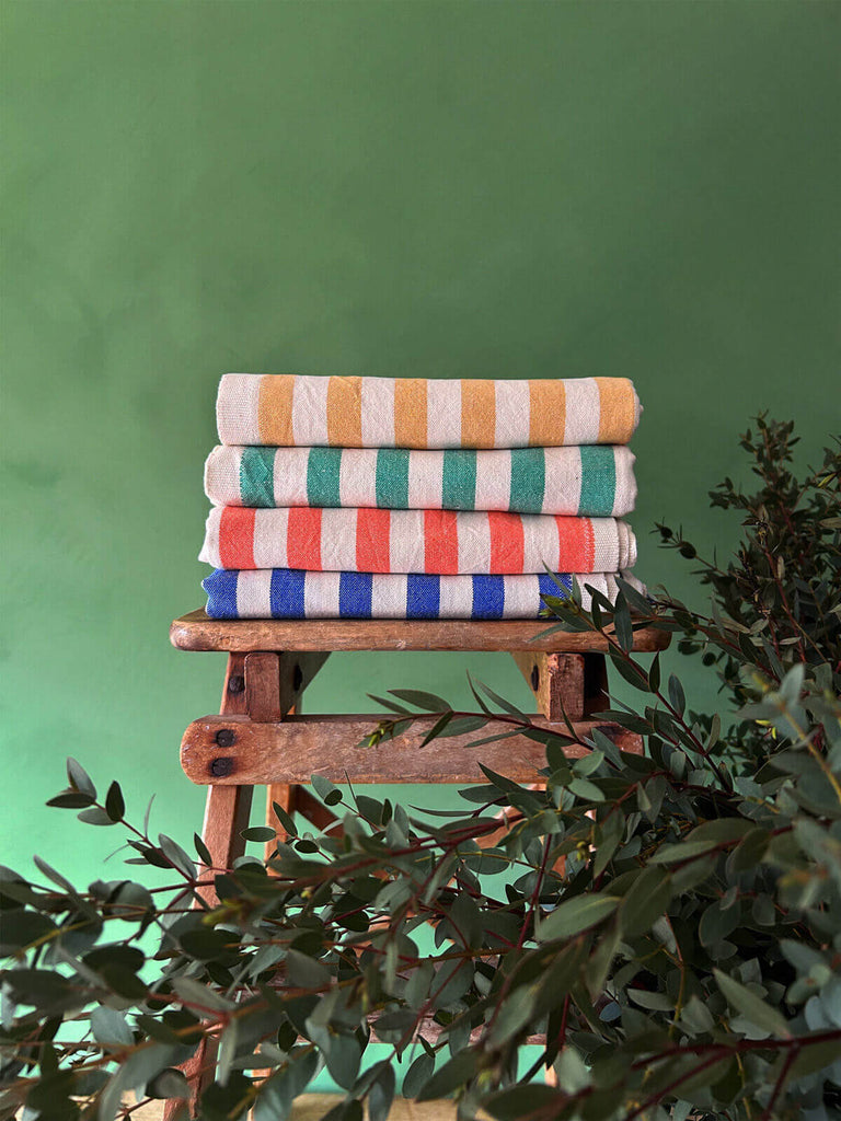 Stack of Brighton stripe hammam towels with colourful holiday-inspired stripes folded on a wooden stool against a lush green background | Bohemia Design
