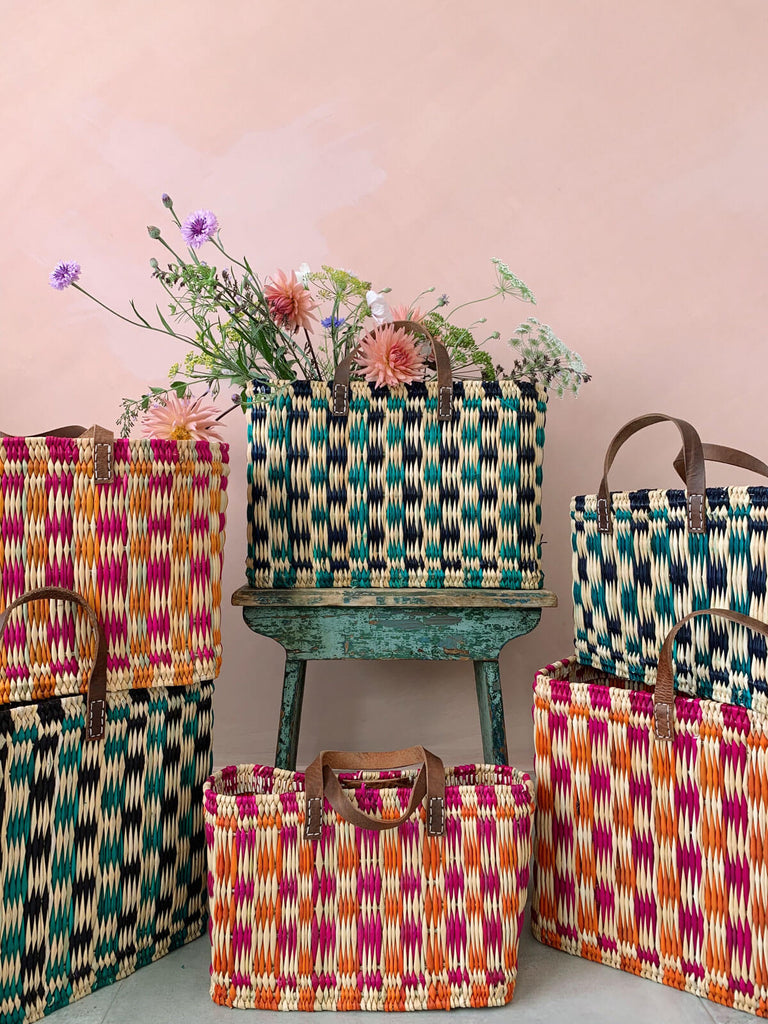 Group of handwoven chequered reed basket bags with leather handles in pink, orange and green in front of a pink wall. One basket filled with flowers.