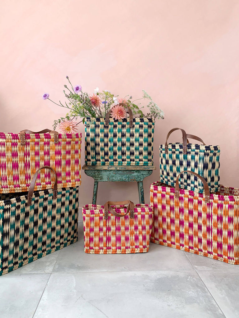 Group of handwoven chequered reed basket bags in pink, orange and green in front of a pink wall. One basket filled with flowers.