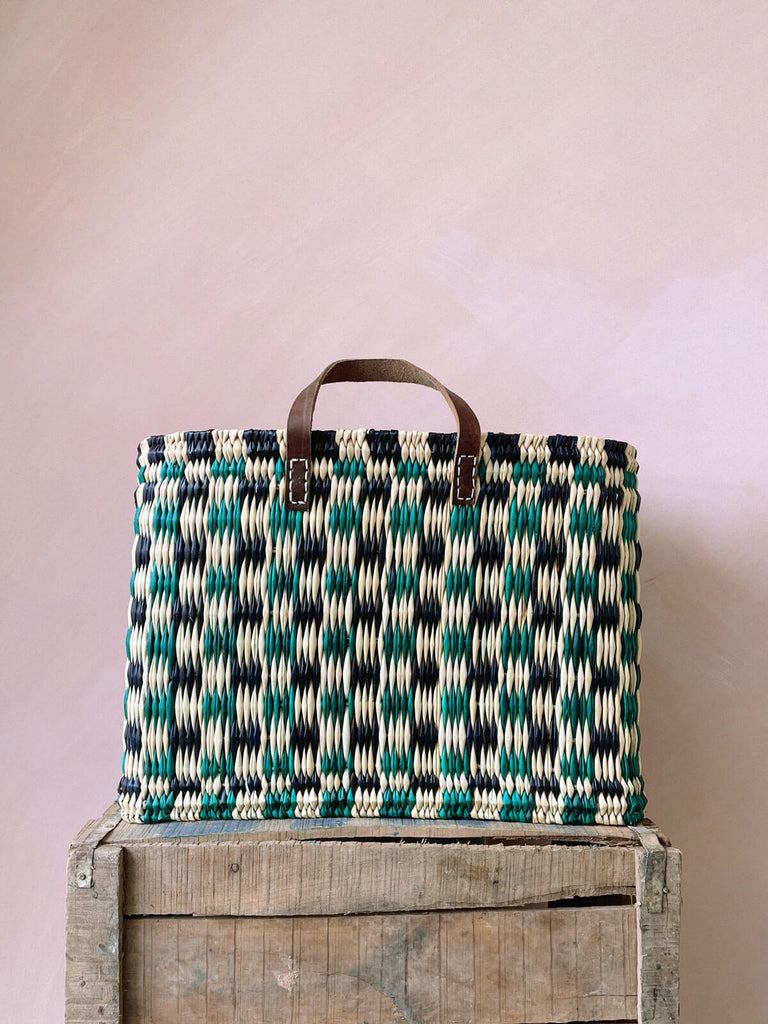 A large chequered green and indigo woven reed basket bag with leather handles on a wooden crate