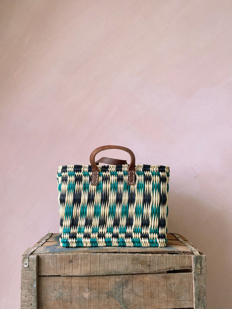 A small chequered green and indigo woven reed basket bag with leather handles on a wooden crate in front of a pink plaster wall