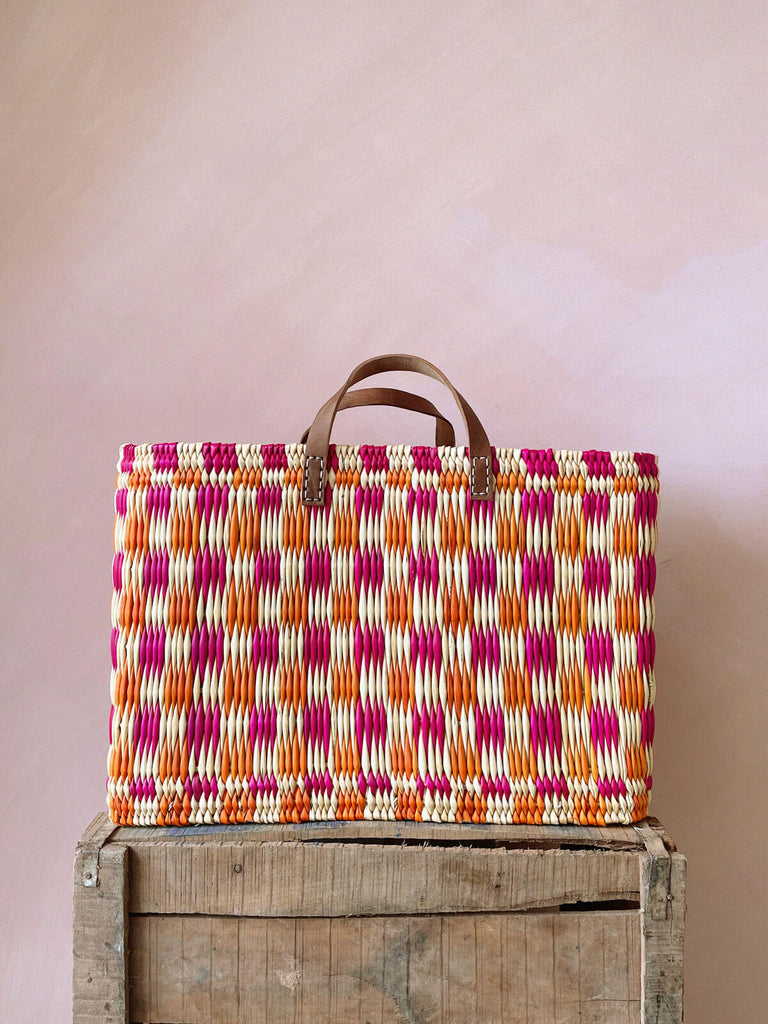 A large rectangular pink and orange chequered woven basket bag sitting on a wooden crate