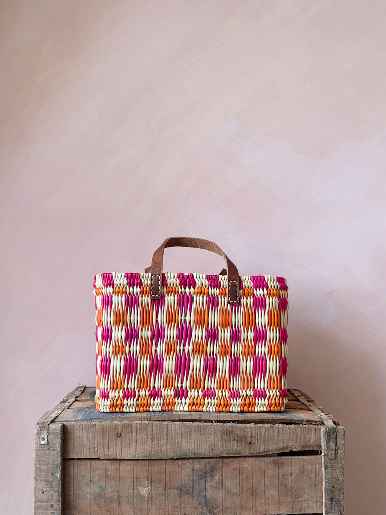 A small, rectangular, pink and orange chequered woven reed basket bag with leather handles on a wooden crate