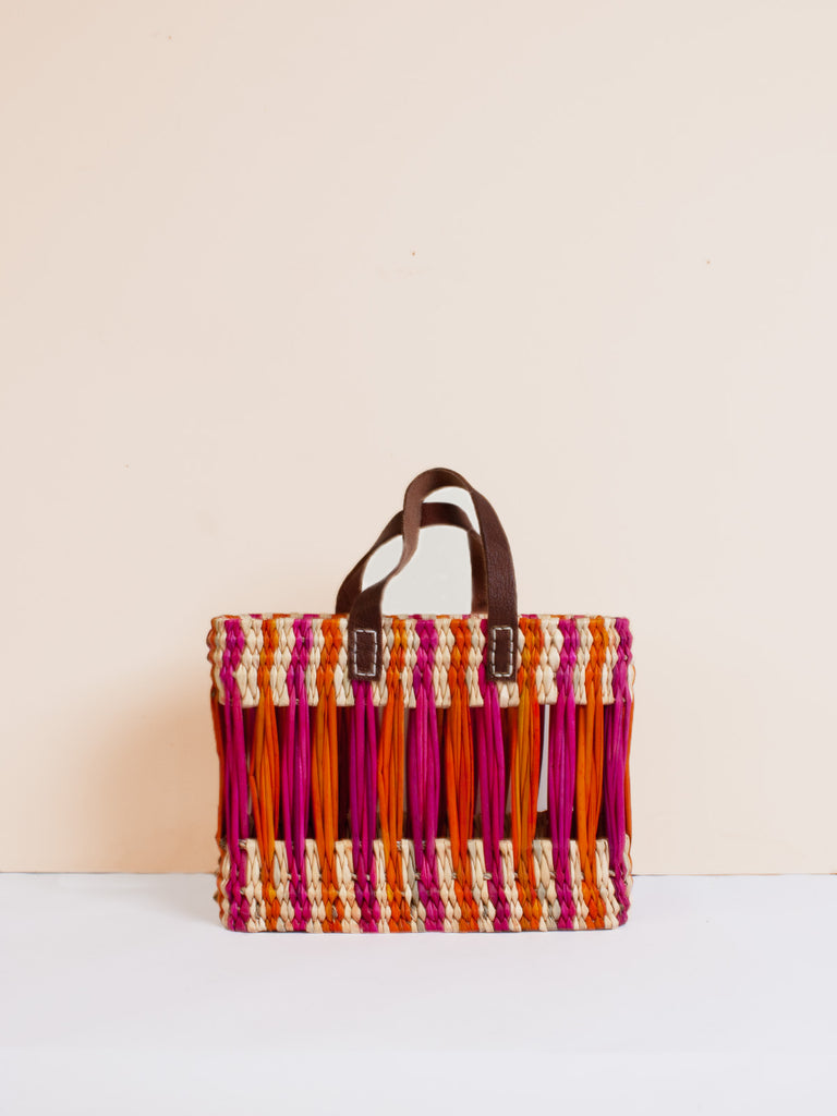 Small boxy woven reed basket bag with decorative pink and orange stripes and leather handles