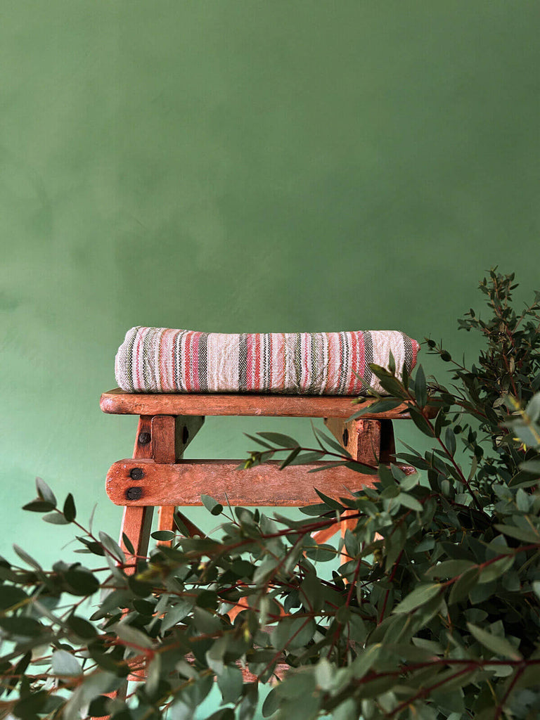Pink and terracotta striped cotton hammam towel on a rustic wood stool with greenery
