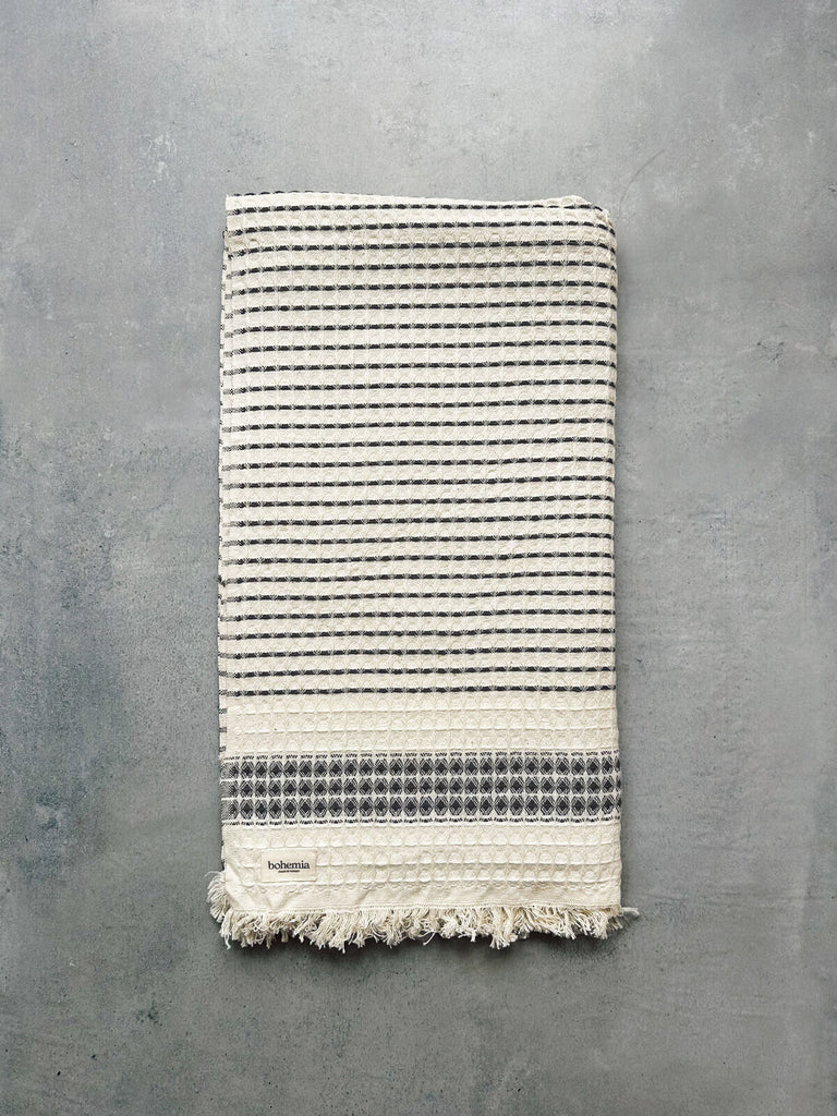 A luxurious and waffle-textured Milos Hammam Towel, showcasing a charcoal stripe and hand-tied tassel fringe, presented against a grey textured background