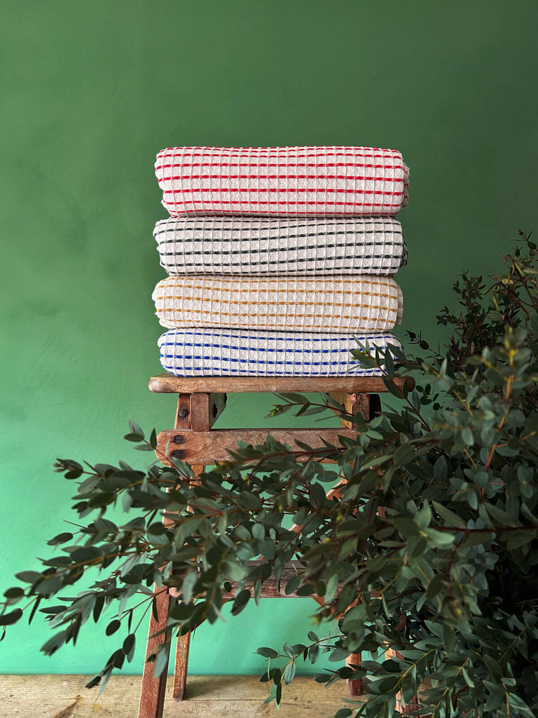 A pile of Milos Hammam Towels in four distinct colours, highlighting the beautiful soft waffle texture of the cotton fabric against a backdrop of lush green wall and foliage