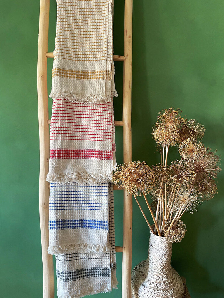A display of beautifully tactile Milos Hammam Towels in four distinct colourways, presented on a rustic wooden ladder against a deep, vibrant green wall | Bohemia Design