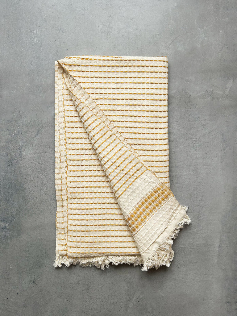 A luxurious Milos Hammam Towel, showcasing a mustard yellow stripe and hand-tied tassel fringe, presented against a grey textured background