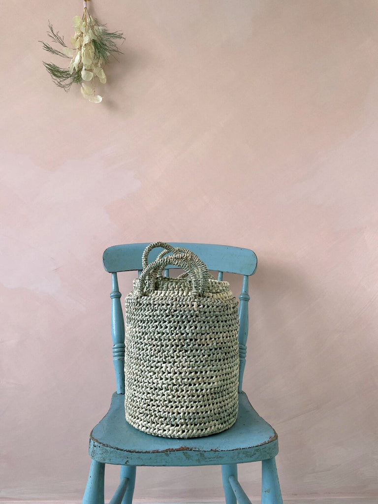 All three nesting baskets stacked inside one another on a blue chair against a pink wall. A posy of dried foliage hangs above.