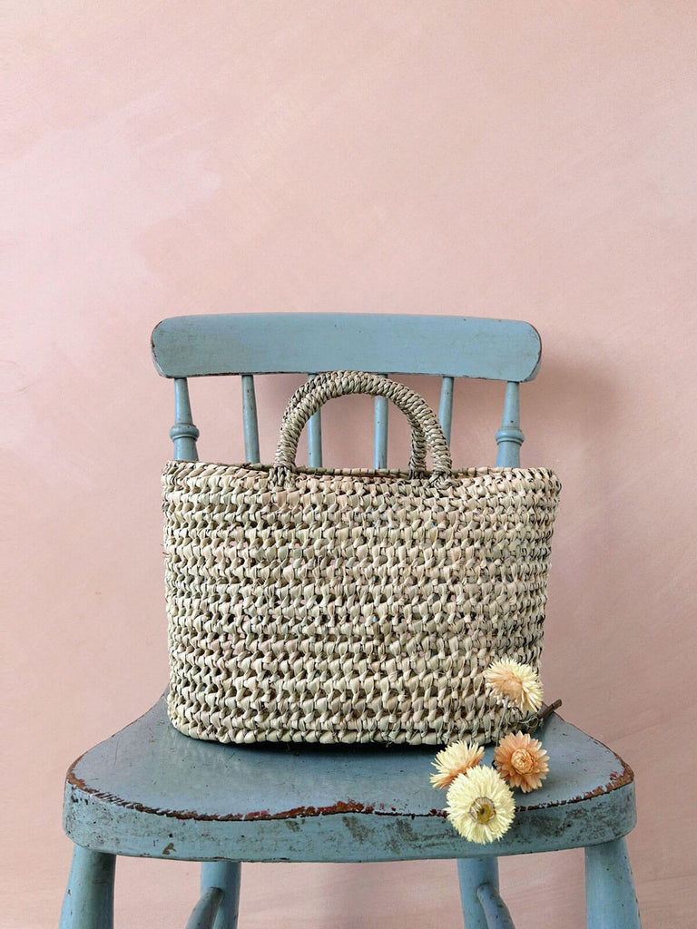 Small oval open weave straw basket with open weave pattern hand woven from natural palm leaf on a rustic blue chair