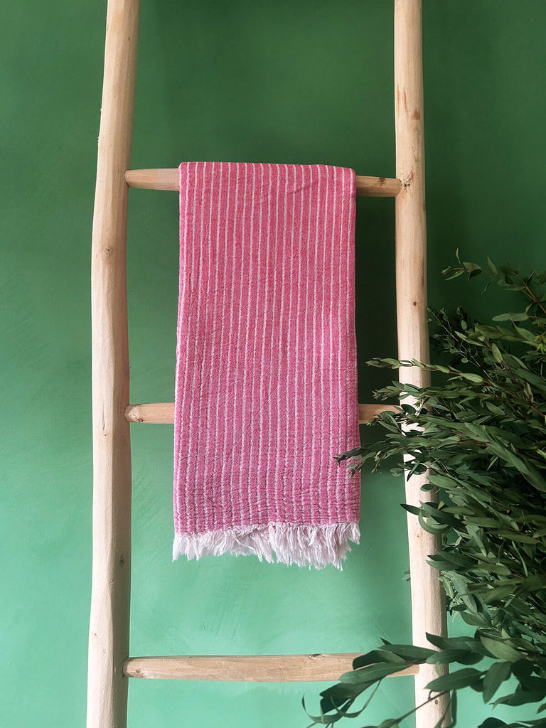 Flamingo pink hammam towel with subtle white stripes, showcased on a rustic wooden ladder against a vibrant green wall | Bohemia Design