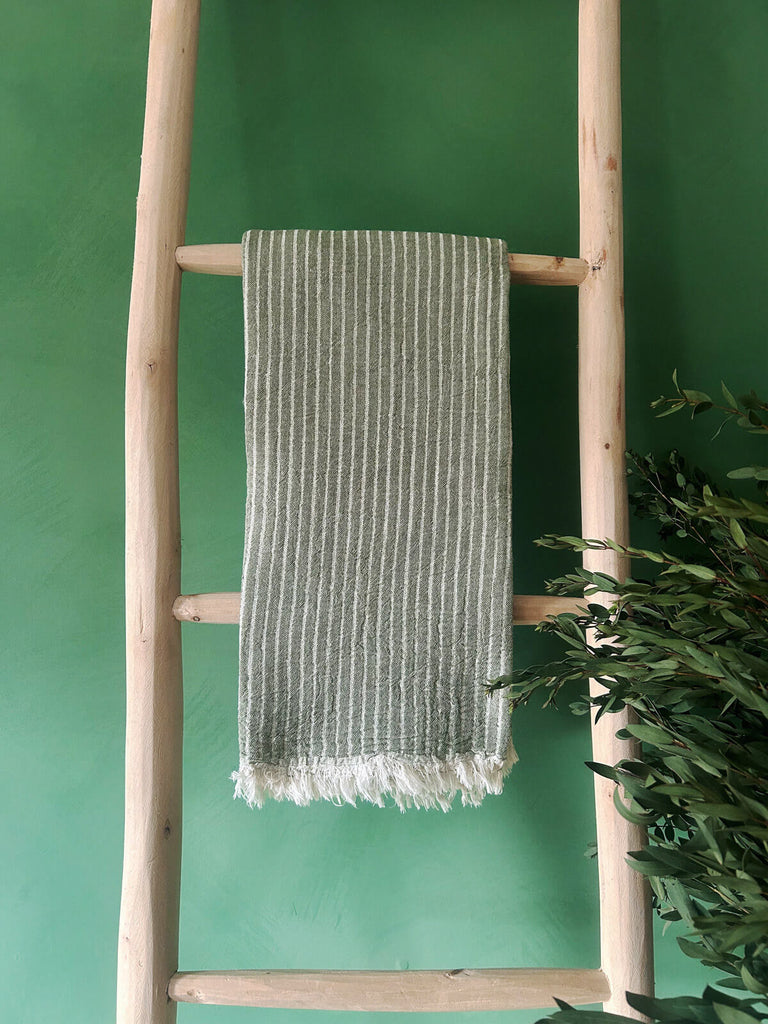 Soft cotton Turkish hammam towel in olive green with subtle woven stripes on a rustic wooden ladder against a vibrant green wall | Bohemia Design