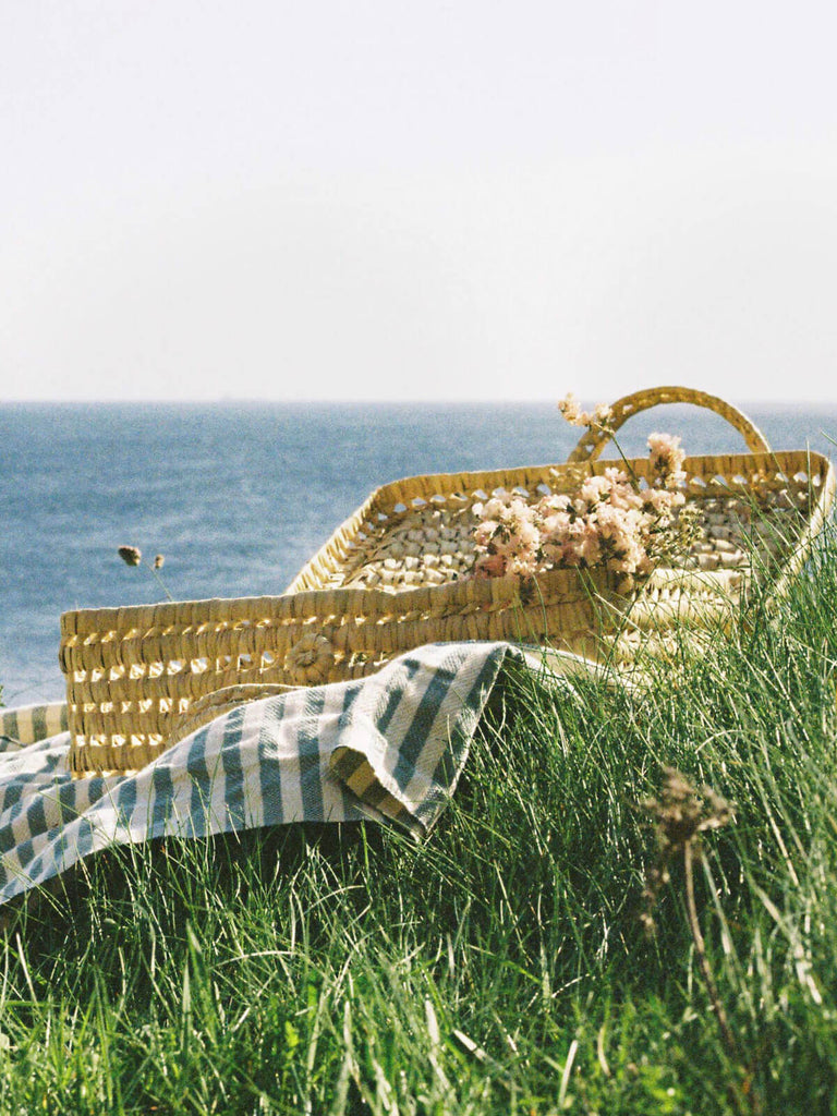 Natural woven suitcase basket on a striped picnic blanket by the coast