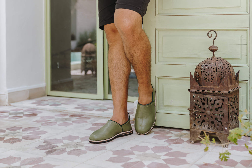 Moroccan berber babouche slippers in olive leather worn by model standing on terracotta tiles with Moroccan lamp