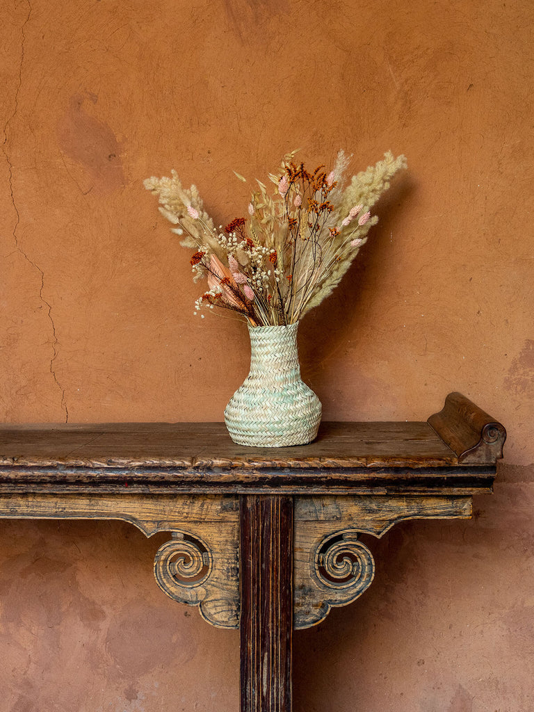 Palm leaf woven vase by bohemia design with dried flowers on a wooden mantlepiece against a terracotta wall