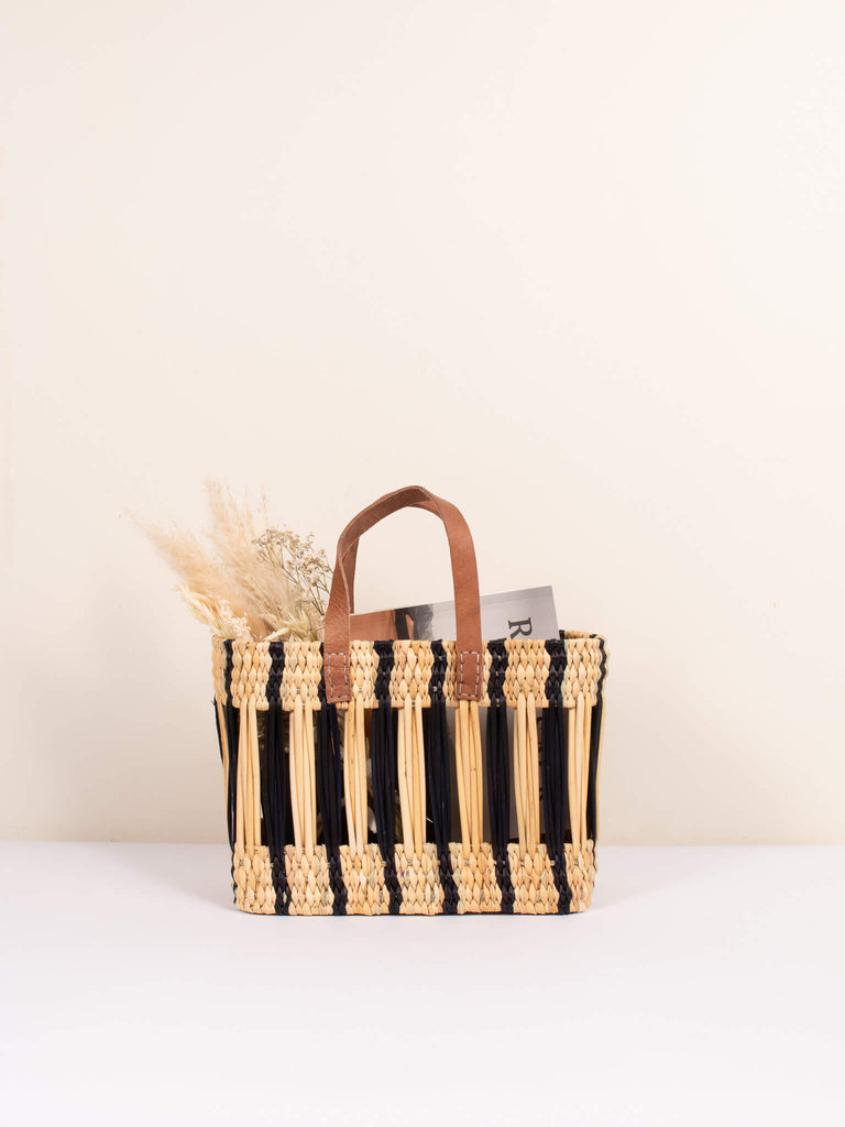 Small decorative reed basket bag with chic indigo stripes and short leather handles holding dried flowers and a travel magazine