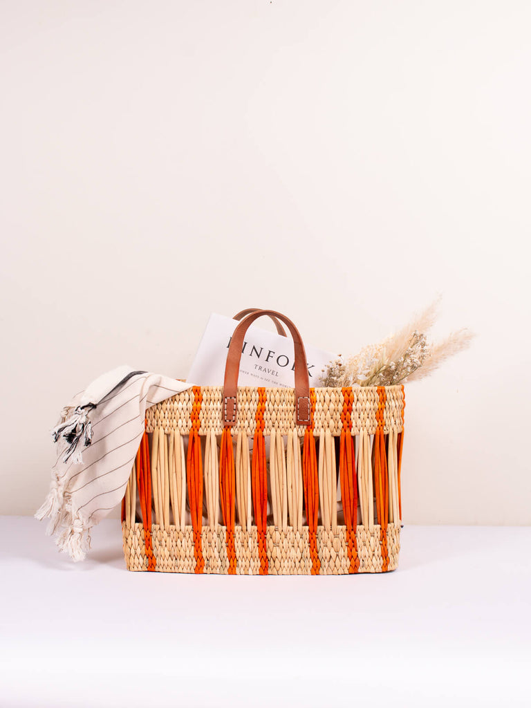 Decorative orange stripe natural woven reed basket bag with dried flowers, travel magazine and scarf