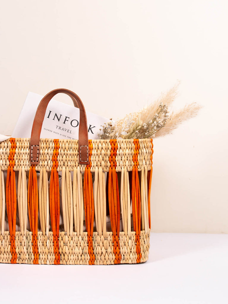 Orange stripe reed woven basket bag with leather handles holding dried flowers and travel magazine