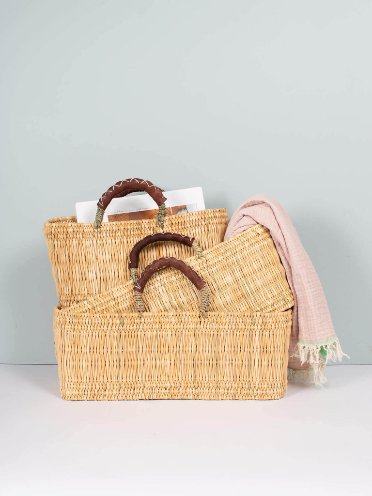 Three sizes of natural woven reed baskets with leather handles- versatile and sustainable for shopping or home storage.
