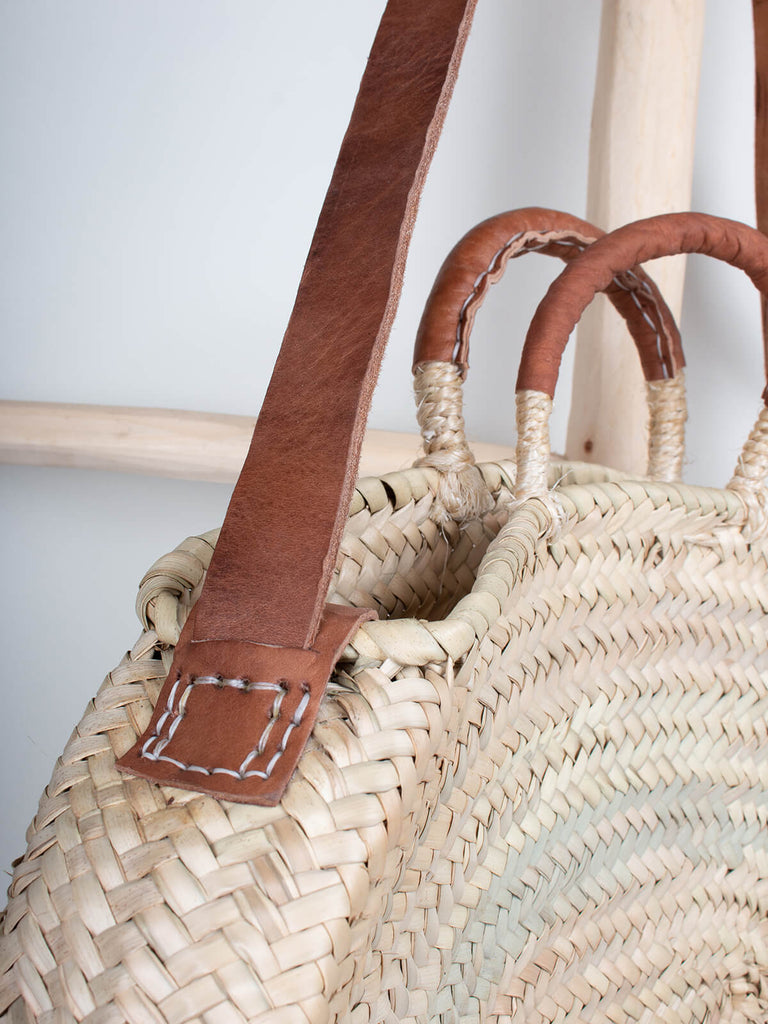 Detail of Moroccan woven basket in an oval shape with tan leather strap and handles hanging from a wooden ladder