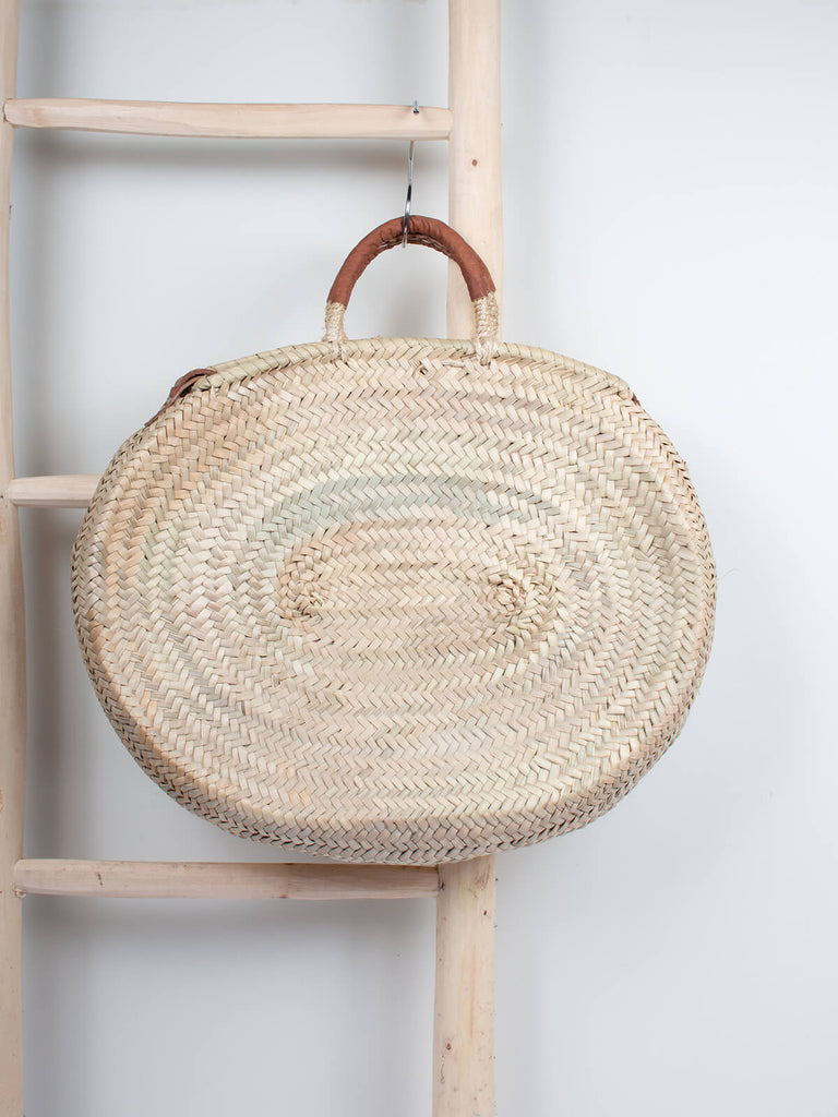 Moroccan woven basket in an oval shape with tan leather strap and handles hanging from a wooden ladder